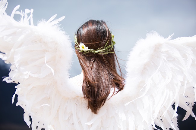 A brunette woman stands with her back to the camera, with a flower garland in her hair and white angel wings on her back. Will we be angels in Heaven? Randy Alcorn says no.