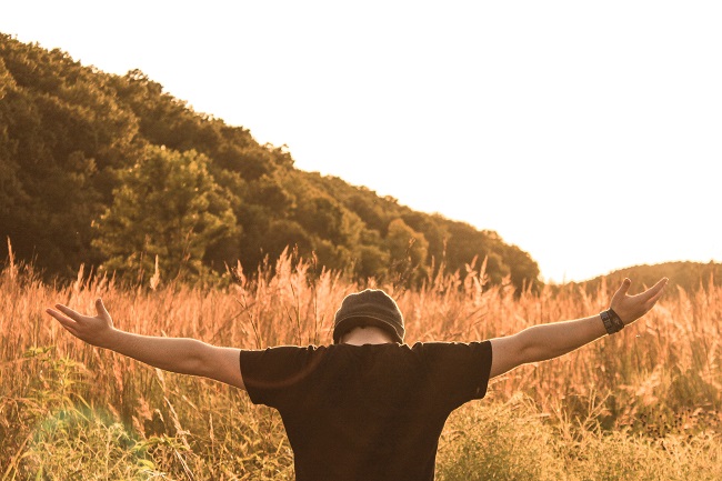 A young man in a t-shirt and beanie hat stands with his back to us, with his arms open wide and his palms facing upwards, he is in prayer.