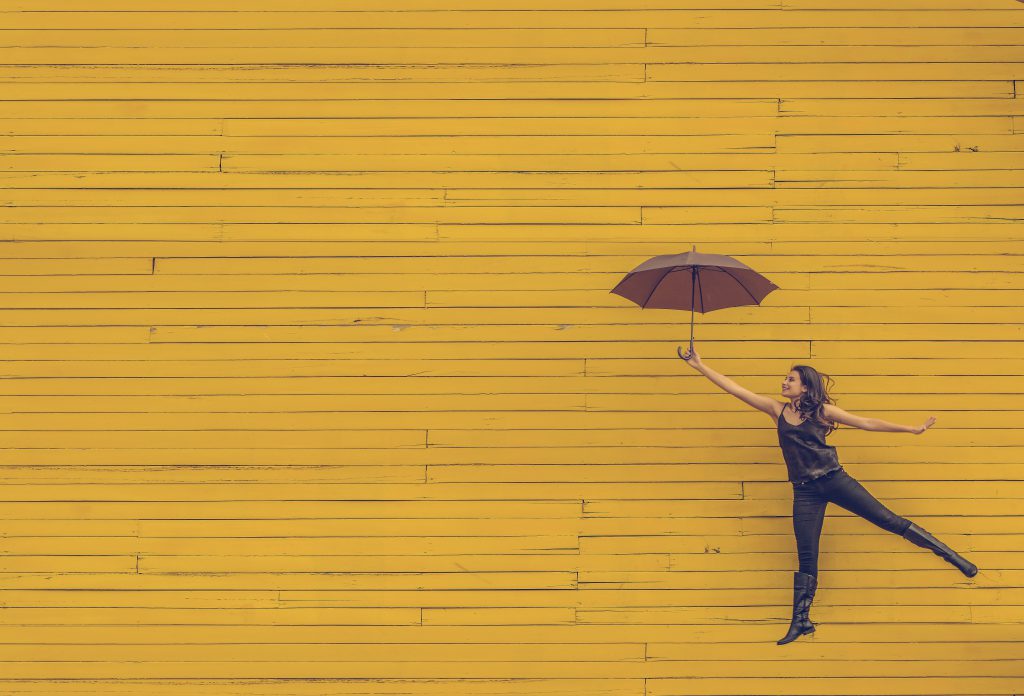 Woman jumping for joy with an open umbrella, in front of a yellow wall. best devotionals for new believers, 60 days to Happiness devotional. Woman jumping for joy with an open umbrella, in front of a yellow wall. best devotionals for new believers, 60 days to Happiness devotional.
