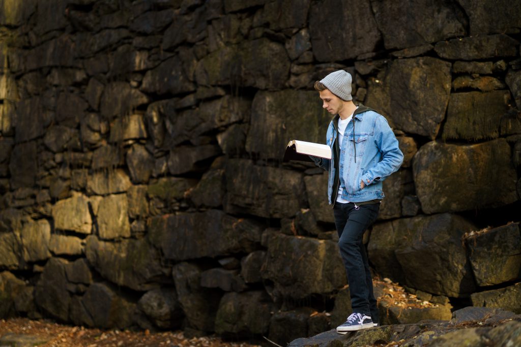 A young man with a denim jacket and beanie reads the Bible in front of an old stone wall. He loves his Bible study. A young man with a denim jacket and beanie reads the Bible in front of an old stone wall. He loves his Bible study.