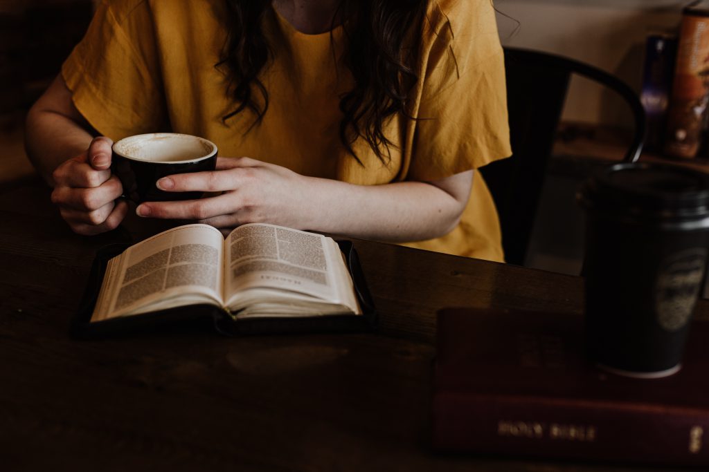 A lady in a yellow top and coffee in her hands reads her Bible study on top of a wooden table. A lady in a yellow top and coffee in her hands reads her Bible study on top of a wooden table.