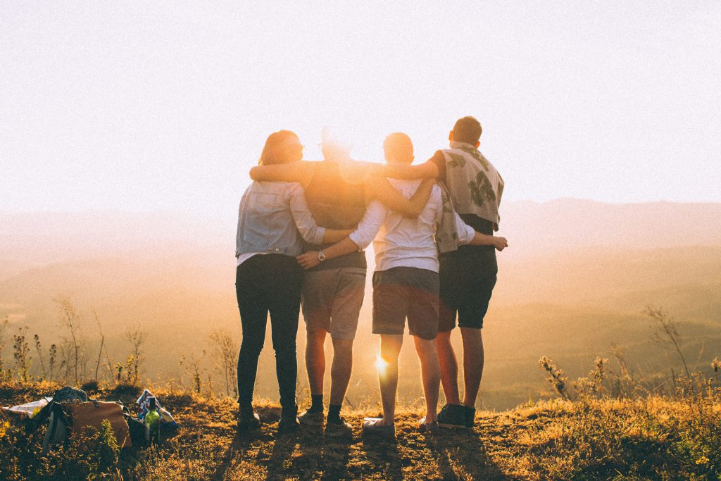Four small group friends stand together on top of a hill, watching the sunset. Four small group friends stand together on top of a hill, watching the sunset.