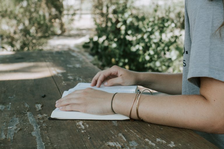 A young woman with bangles on her right wrist, sits at a wooden table in a yard reading her Bible. Want to know where to start reading the Bible? Start with the Gospel of John!